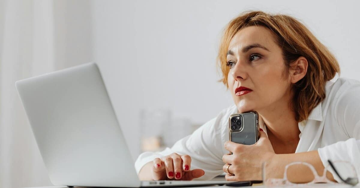 serious woman holding phone while working on laptop